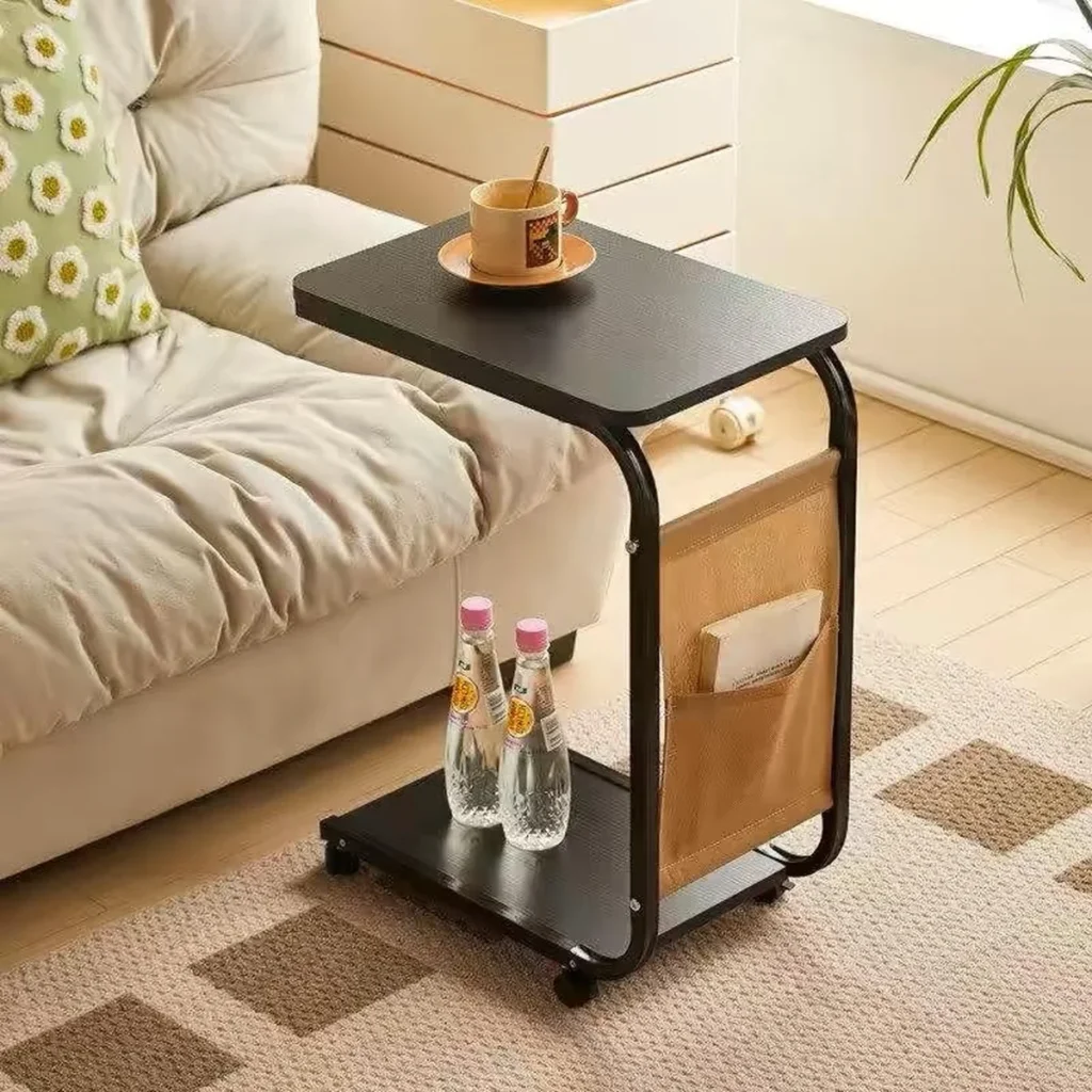 Portable laptop table with a black tabletop, metal frame, storage pouch, and bottom shelf holding two water bottles, placed next to a cozy beige sofa in a modern living room.