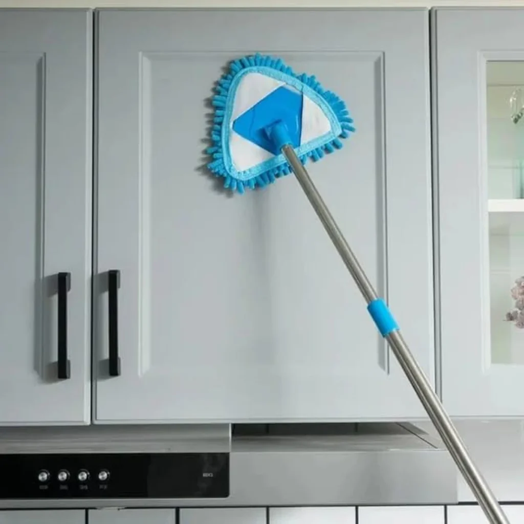 Microfiber triangle-shaped mop cleaning a gray kitchen cabinet. The mop features a blue and white washable microfiber pad with a stainless steel extendable handle, ideal for dusting and deep cleaning surfaces.