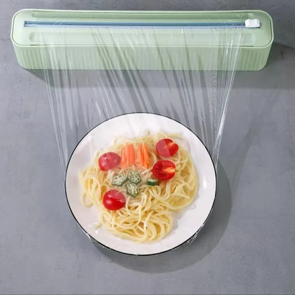 Magnetic foil and cling wrap dispenser mounted on a wall, dispensing transparent food wrap over a plate of spaghetti with cherry tomatoes, okra, and carrots.