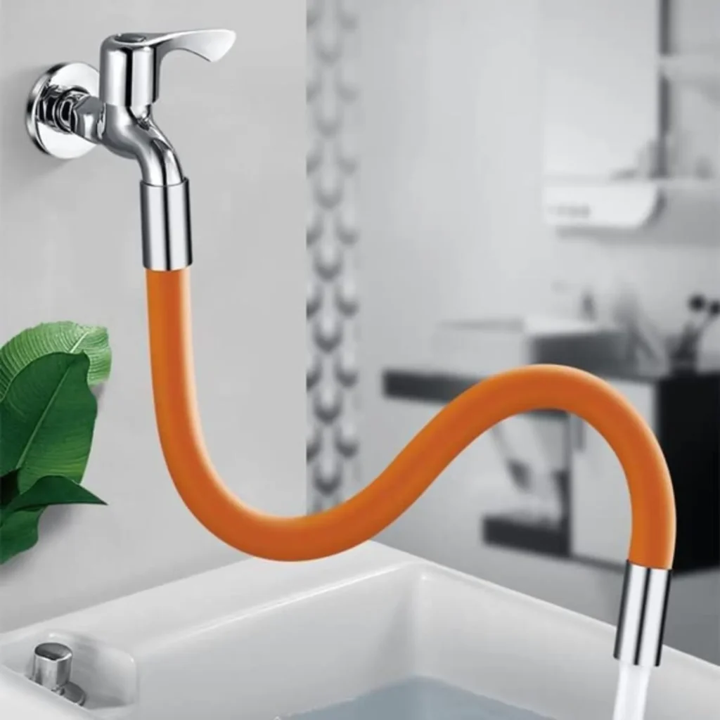 A flexible, orange silicone faucet extender attached to a wall-mounted chrome tap. The nozzle pipe is shapeable, bending into an S-curve, directing water into a white sink. A green plant leaf is visible in the corner, with a blurred modern bathroom setting in the background.